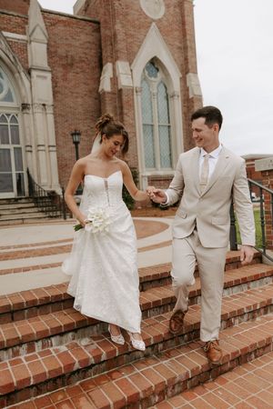 Smiling newlyweds descend brick steps outside historic church, bride in strapless lace gown holding white bouquet.