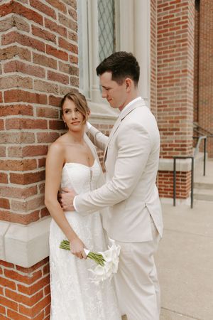 Groom in cream suit embraces bride in lace gown against red brick church wall with bouquet.