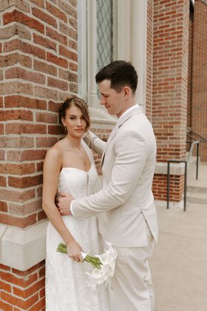 Bride and groom pose against brick church wall, she holds white calla lily bouquet in lace gown, he wears ivory suit.