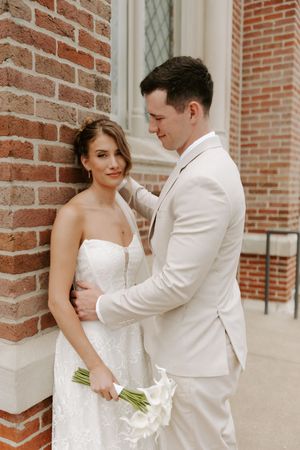 Bride in strapless lace gown holds calla lilies as groom in ivory suit embraces her against brick church wall.