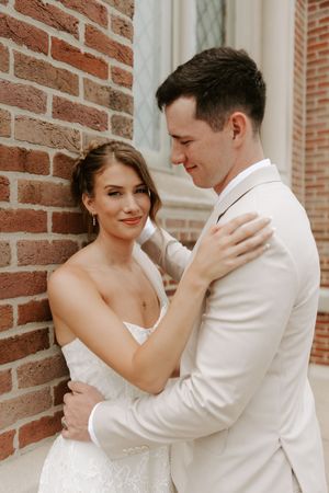 Bride and groom embrace tenderly against a brick wall, she in a lace gown, he in a white suit.