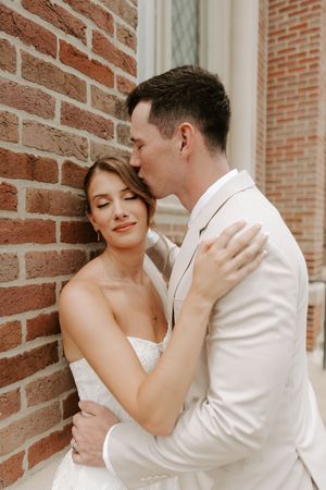 Groom tenderly kisses bride on temple as she closes her eyes against red brick church wall.
