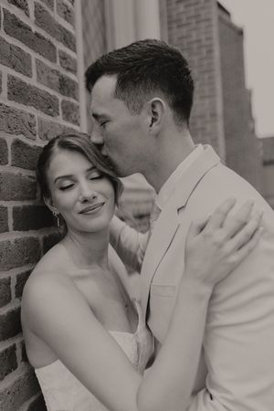 Black and white photo of a groom in a white suit kissing his bride on the temple against a brick wall.