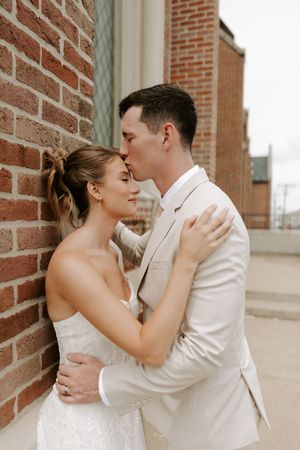 Groom in beige suit kisses bride in strapless lace gown against a rustic red brick wall outdoors.