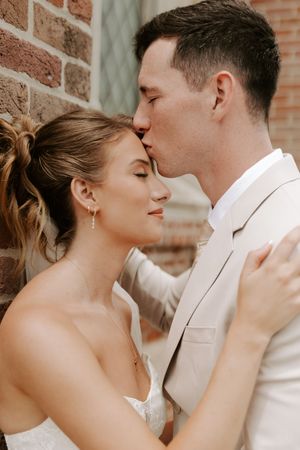 Groom in tan suit tenderly kisses bride on forehead against a rustic brick wall on their wedding day.
