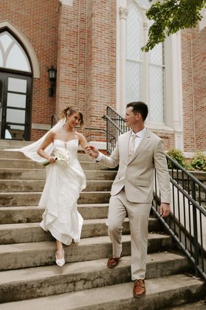 Bride and groom descend church steps hand in hand, bride in strapless white gown, groom in tan suit.