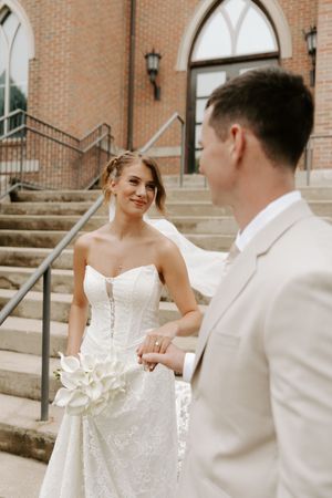 Bride in strapless lace gown holding white bouquet smiles at groom on steps outside a brick church building.