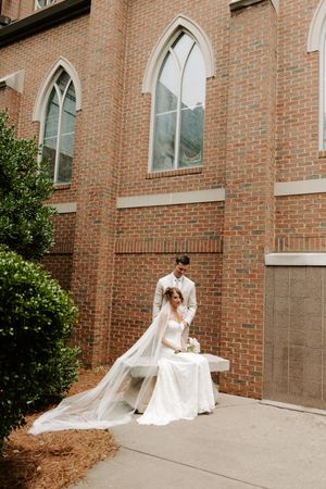 Bride in flowing veil sits on bench beside groom outside a brick church with arched windows.