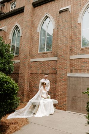Bride seated outside brick church with gothic windows, groom standing behind her, veil flowing.