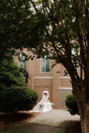 Wedding couple embraces on pathway surrounded by lush green trees outside brick church, bride's veil flows gracefully.