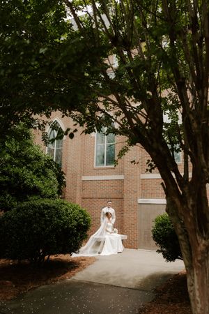 Bride and groom share a romantic moment on a bench surrounded by lush greenery outside a brick wedding venue.