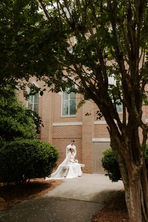 Bride and groom share an intimate moment on a path framed by lush trees outside a brick church.