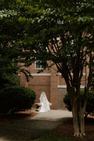 Bride in white gown and veil sitting alone on bench beside brick building framed by green trees.