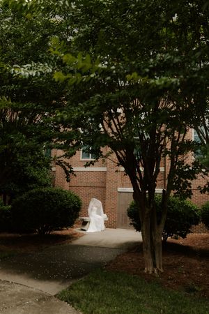 Bride in white wedding dress and veil standing on a brick building pathway surrounded by lush green trees.