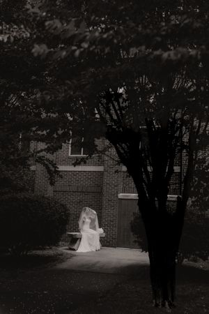 Ethereal black and white photo of a bride in white gown and veil seated near a brick building.