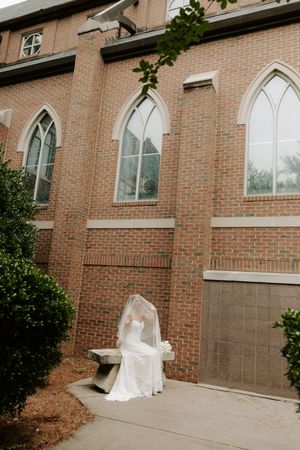 Bride in strapless lace gown and long veil sits on stone bench outside Gothic-windowed brick church.