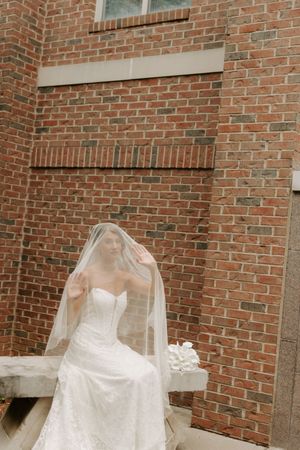 Bride in strapless lace gown with flowing veil seated against red brick wall holding white bouquet.