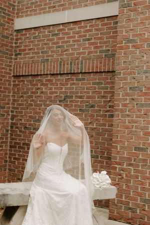 Bride in strapless lace wedding gown sits beneath flowing veil against a red brick wall holding white bouquet.