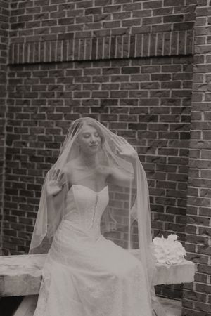 Dramatic black and white portrait of a bride seated against a brick wall with her veil draped overhead.