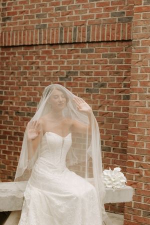 Bride in strapless lace gown lifts sheer veil with eyes closed while seated against rustic brick wall.