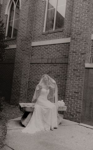Black and white photo of bride in wedding gown seated outside brick church.