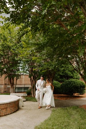 Bride and groom walk hand in hand along garden path surrounded by lush green trees.