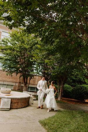 Bride and groom walk hand in hand past a fountain under green trees outside a brick building.