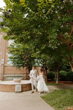 Bride and groom stroll hand-in-hand past a brick fountain surrounded by tall green trees on their wedding day.