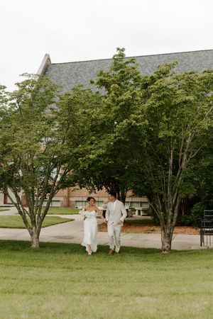 Bride and groom walk hand in hand across a green lawn with trees and a large brick building behind them.