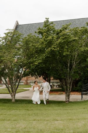 Bride and groom walk hand-in-hand across green lawn with trees and brick building backdrop.