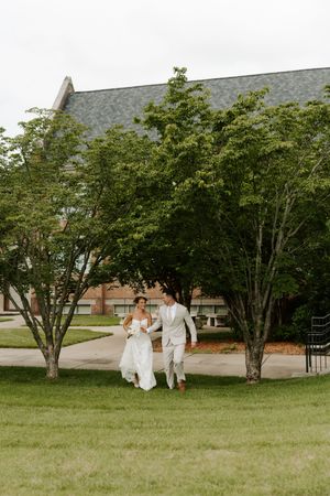 Happy couple in wedding attire strolling through lush garden grounds near elegant brick building.