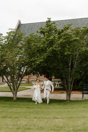 Bride and groom holding hands running across green lawn with large trees and brick building backdrop.