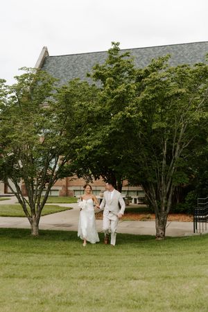 Bride and groom stroll together across a grassy lawn flanked by green trees with a brick building backdrop.