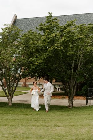 Bride and groom walking hand in hand across green lawn near brick building.