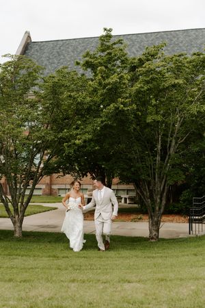 Smiling bride in white gown and groom in tan suit walk together on sunny garden path.