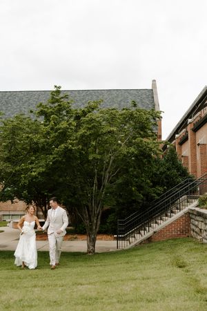 Bride and groom walk hand in hand on a grassy lawn beside a brick building with an iron staircase.