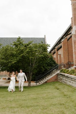 Bride and groom walk hand in hand across green lawn outside brick church on their wedding day.