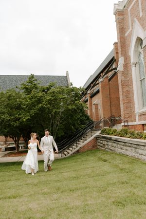 Bride and groom walk across green lawn beside a large brick church on a cloudy day.