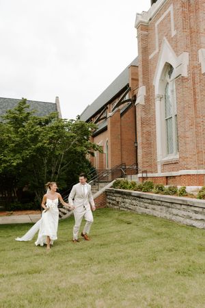 Bride and groom hold hands walking on grass beside ornate red brick church exterior.