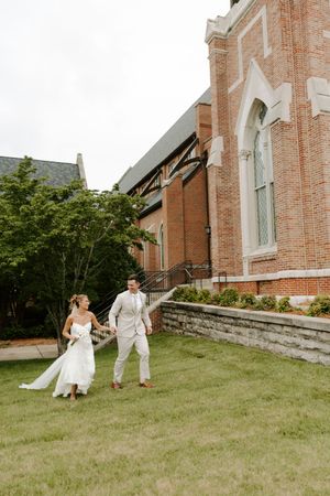 Bride and groom walking together on grass beside historic brick church exterior.
