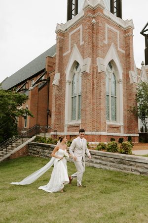 Bride and groom walk hand in hand on the lawn of a Gothic brick church with arched stained glass windows.