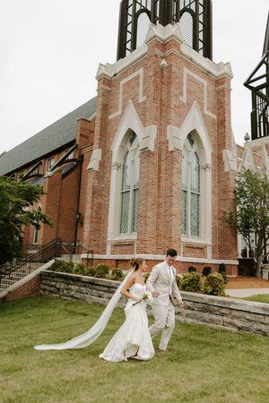 Bride and groom walk together on grass before Gothic brick church tower with arched windows.