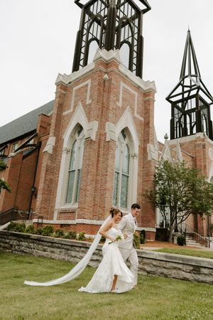 Bride and groom share romantic dip kiss on lawn in front of Gothic brick church steeple.