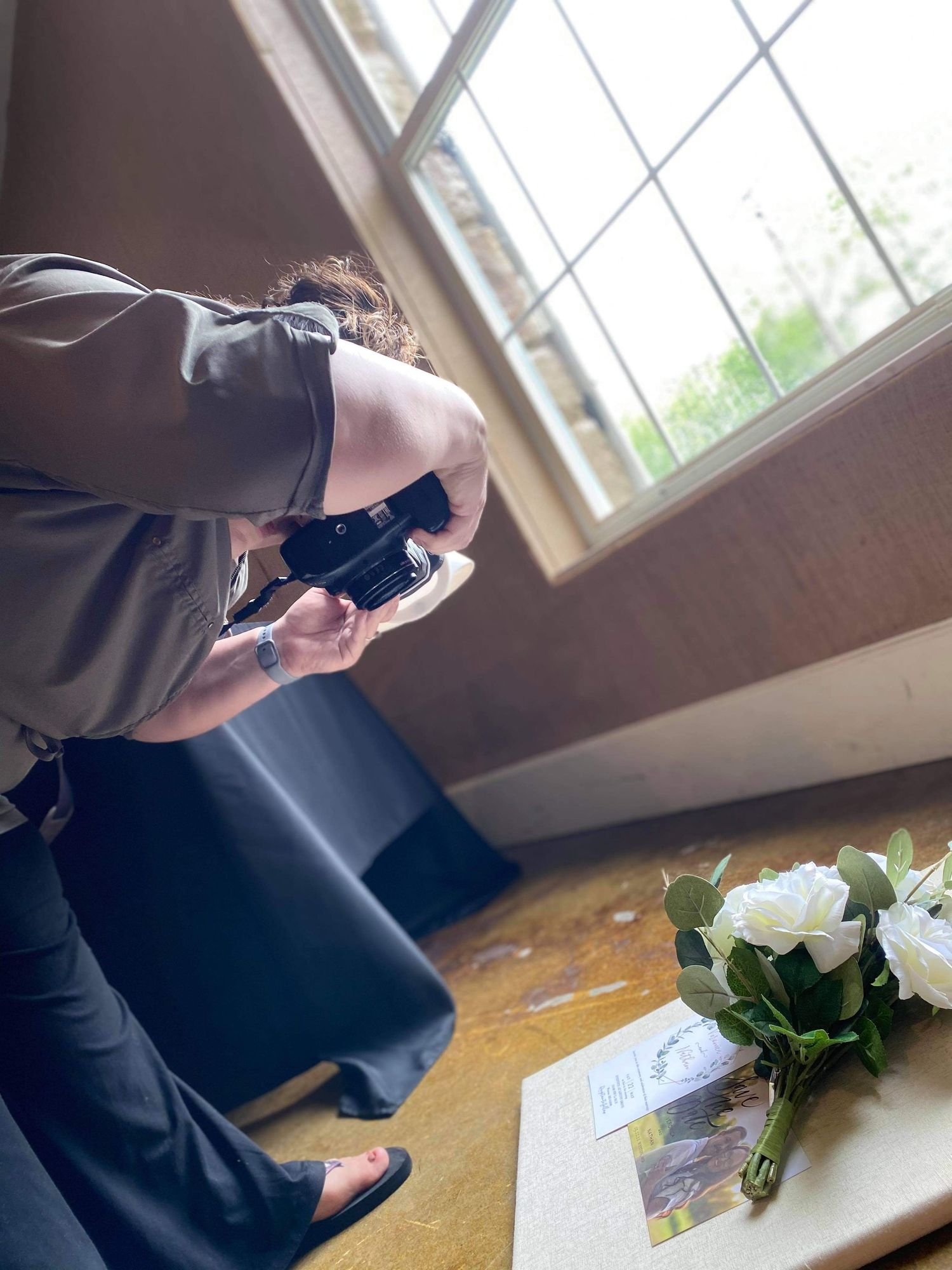 Photographer crouching to capture a white rose bouquet on a wooden table near a window.