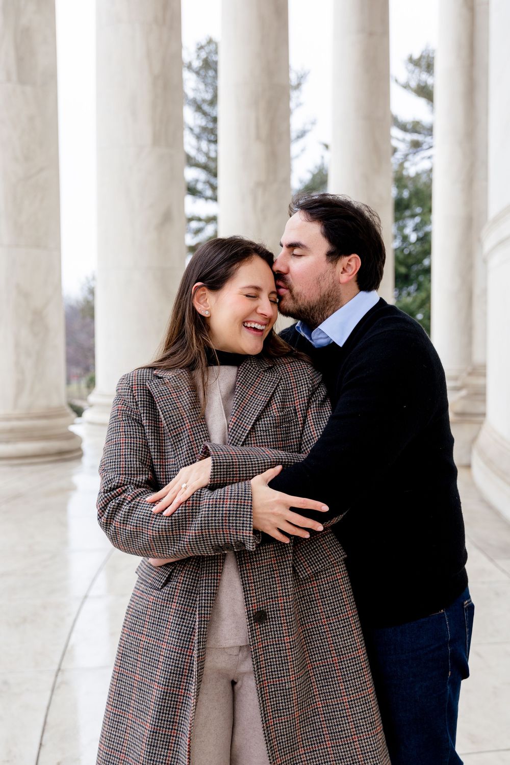 A couple shares a romantic moment at the Jefferson Memorial, with the man kissing the woman's cheek as she smiles joyfully.