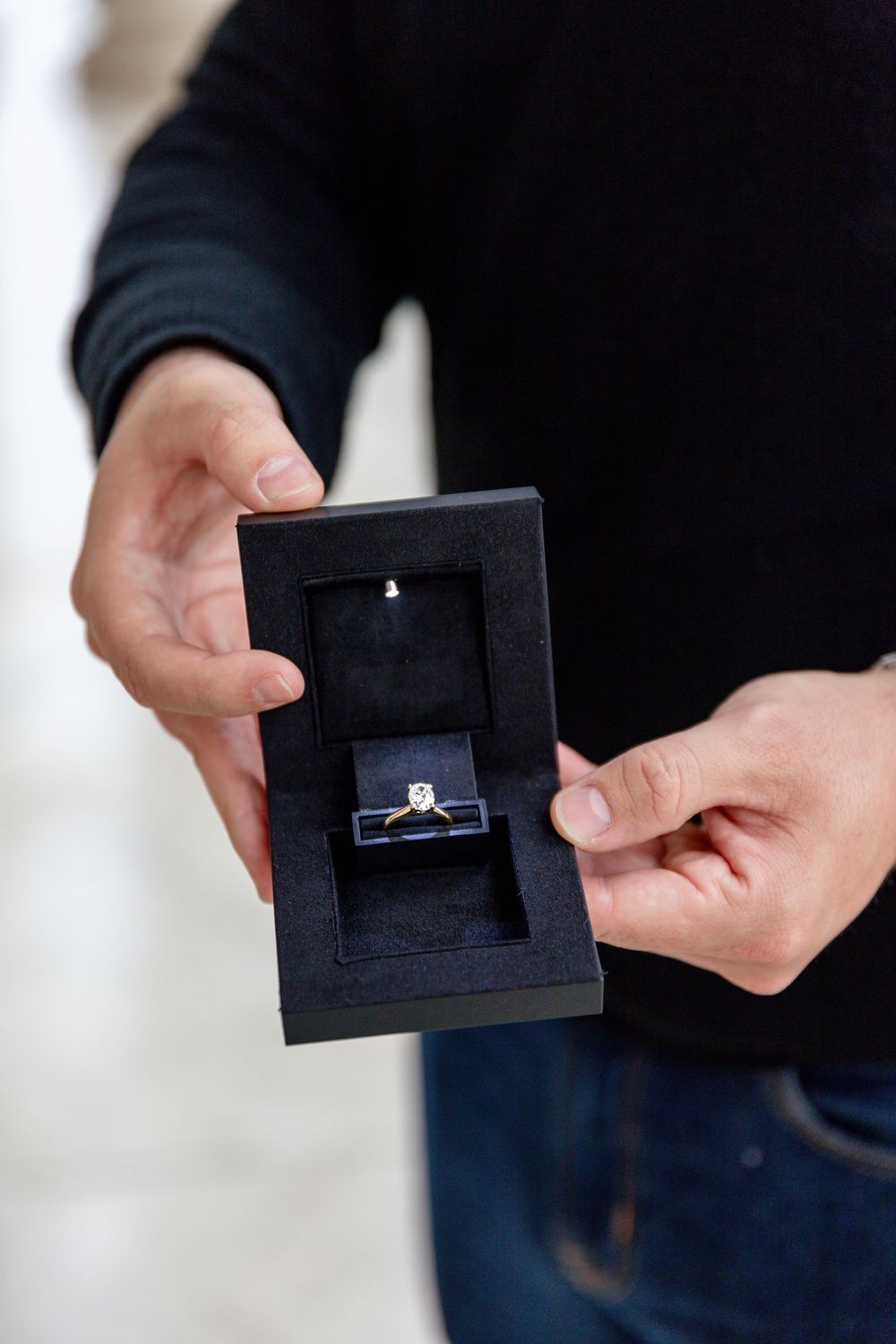 Man in black sweater holding open a dark jewelry box with a diamond solitaire engagement ring inside.