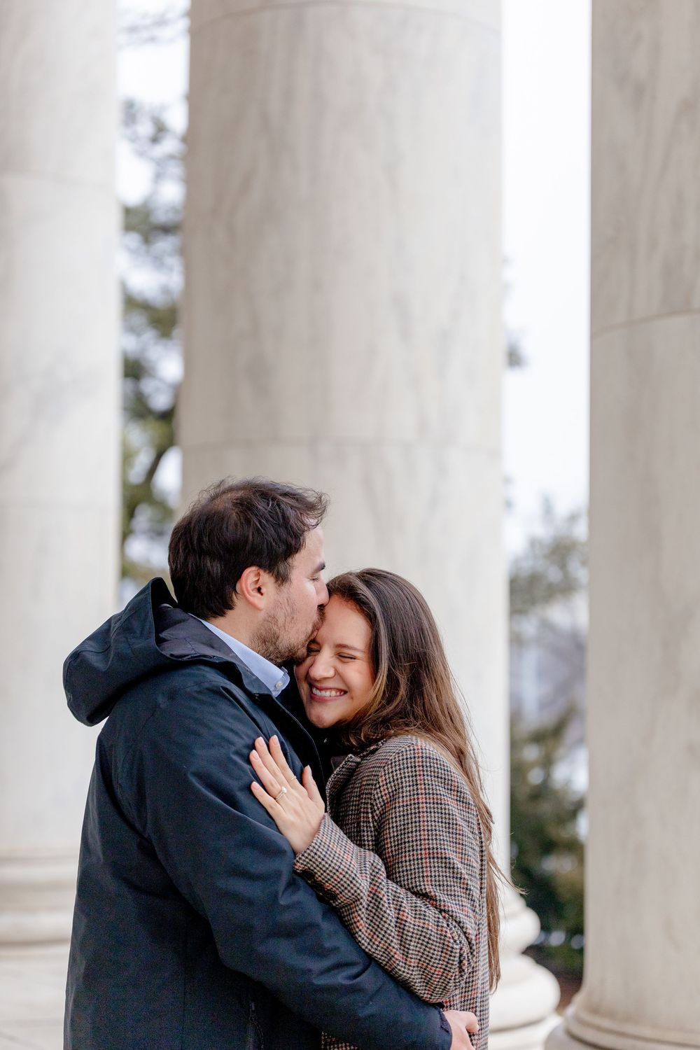 Couple embracing and kissing between marble columns outdoors in winter coats.