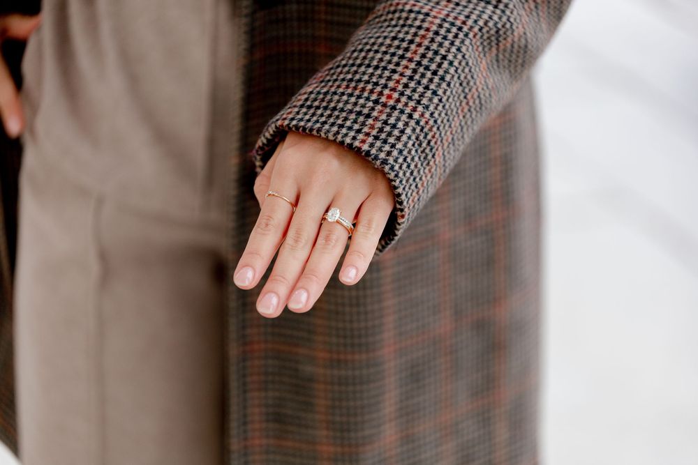 Close-up of a hand wearing a diamond engagement ring, with a plaid coat sleeve visible in the background.