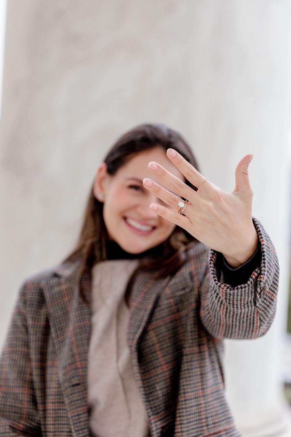 Smiling woman showing off her engagement ring with a gold band while wearing a plaid blazer.