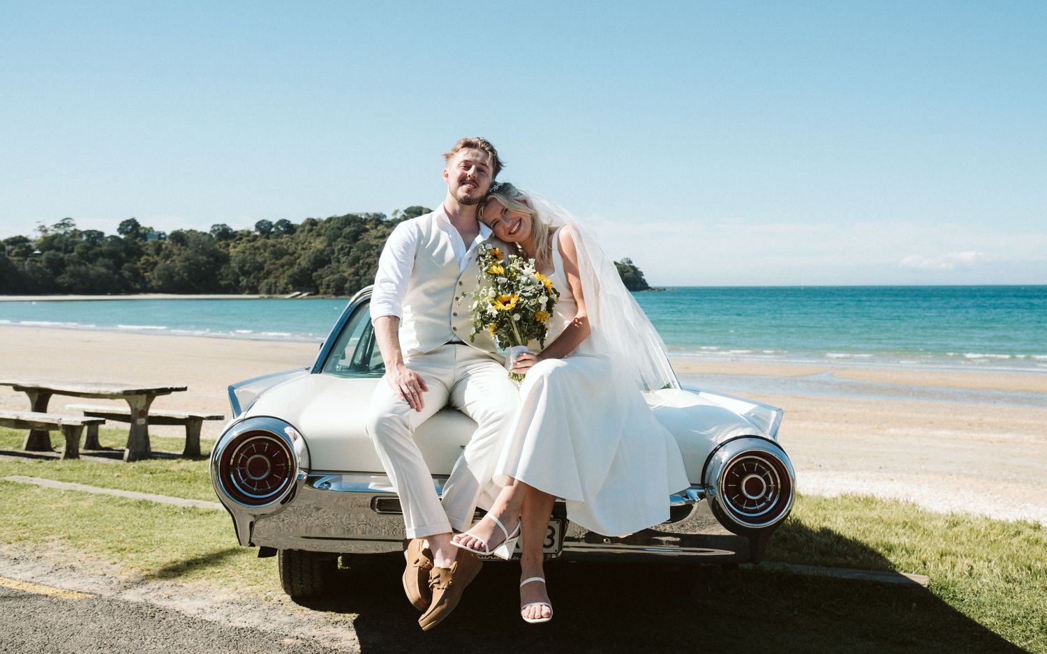 Newlywed couple sitting on a vintage white car at a sunny beach, bride holding sunflowers in wedding attire.